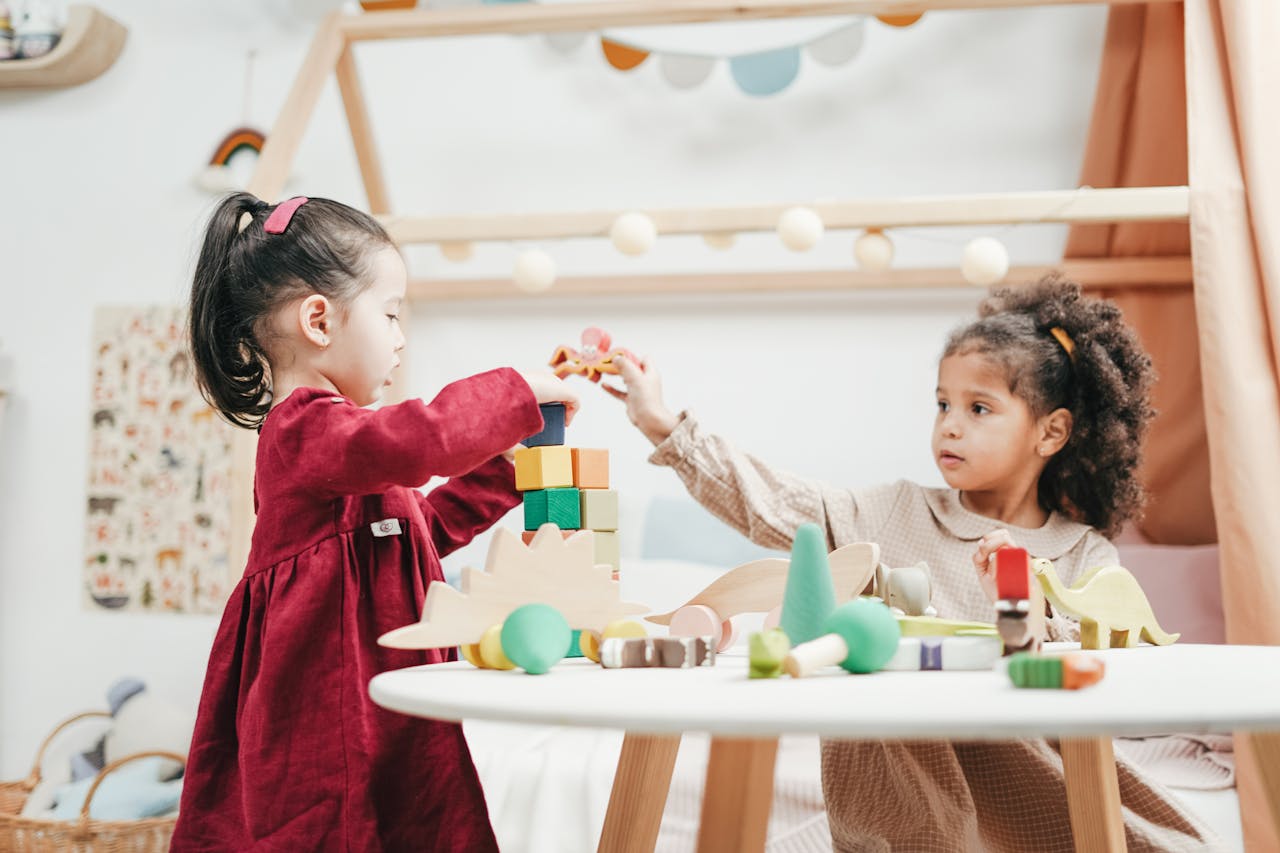 hero Two young girls enjoying playtime with wooden toys indoors in a warm, colorful playroom.